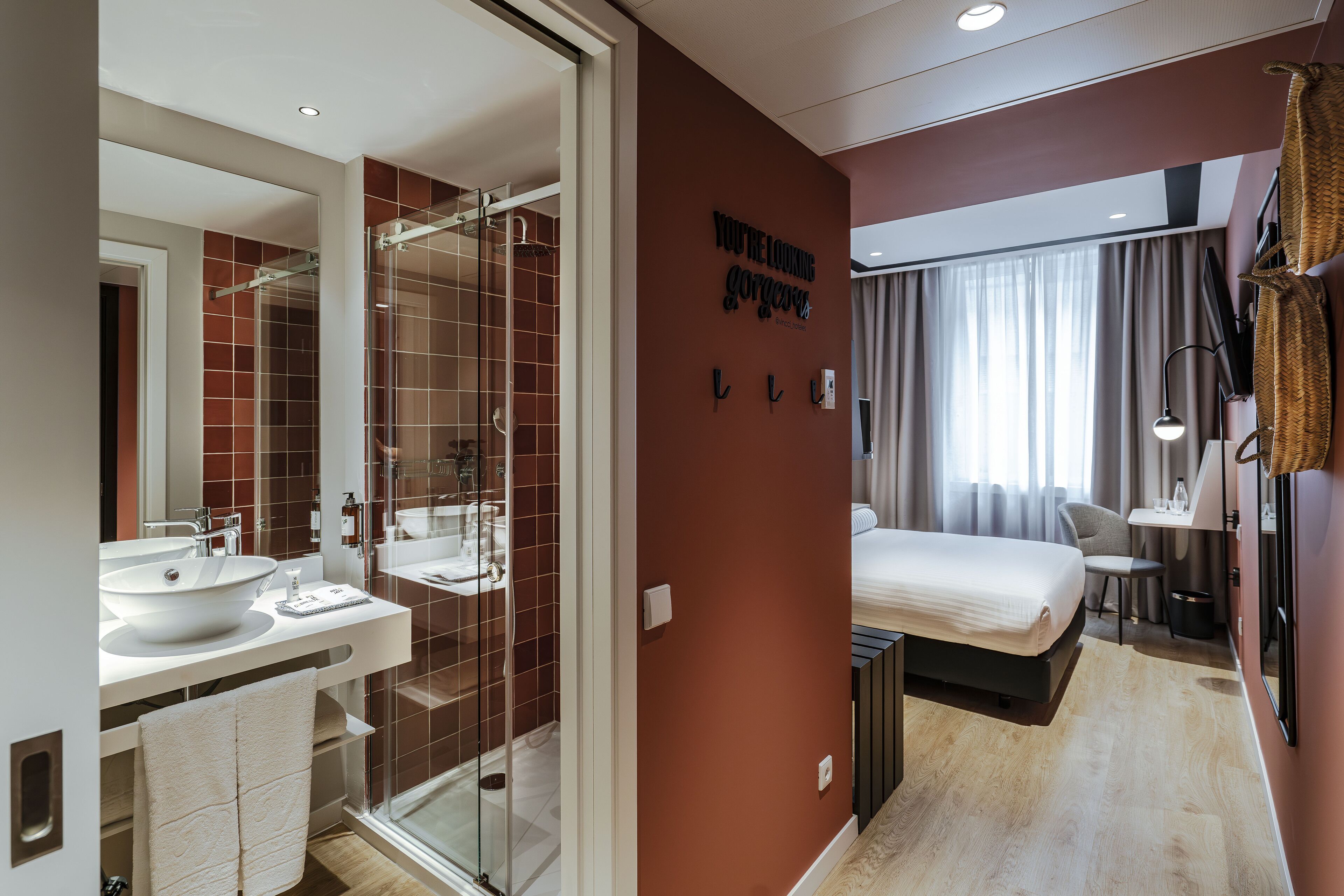 A modern hotel room with a glass shower, a double bed, a desk, and a view of the city.