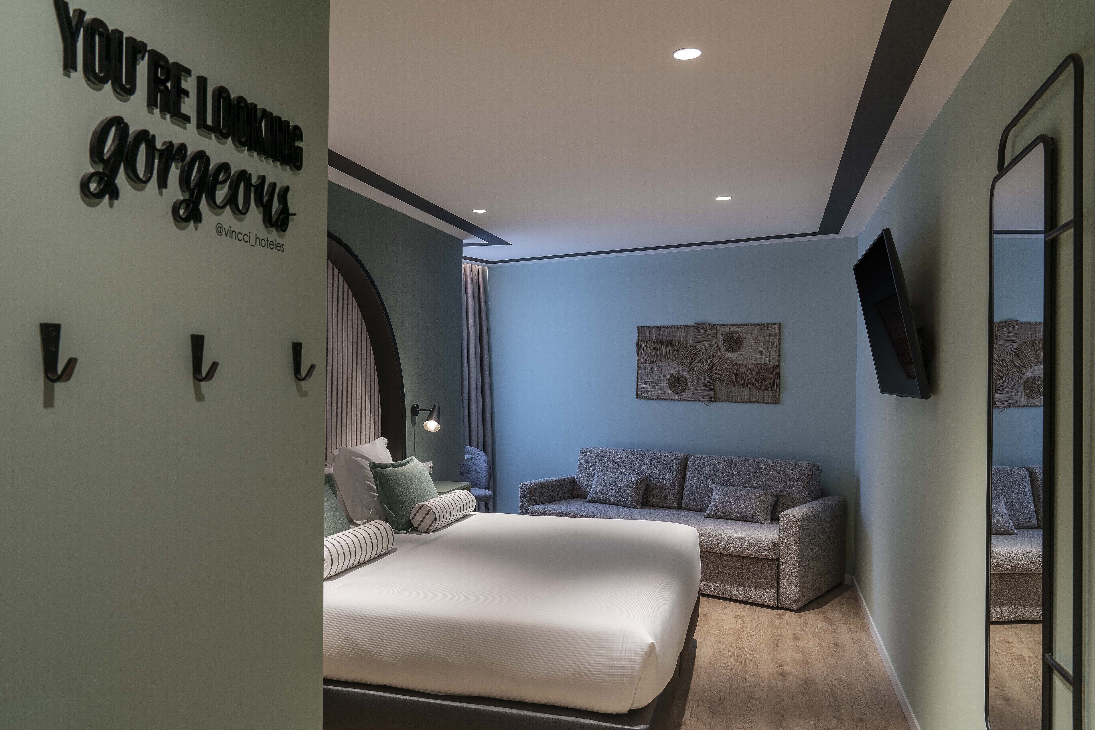 A modern hotel room with a bed, a sofa, a wall-mounted TV, and a decorative mirror.
