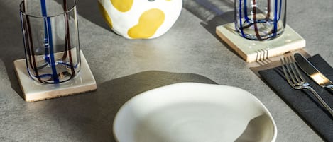 Ein Tisch ist gedeckt mit einem weißen Teller, zwei klaren Gläsern mit blauen Streifen, einer gelb-weißen gepunkteten Vase und einer kleinen Pflanze.