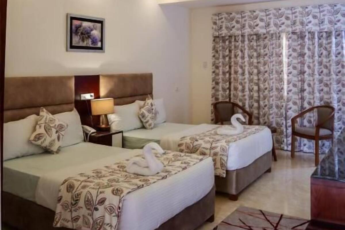 A hotel room with two beds, a nightstand, a lamp, a framed picture on the wall, and a patterned curtain.
