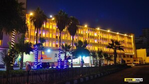 Un hôtel à la façade moderne, illuminée d’une lumière chaleureuse, avec une entrée imposante agrémentée d’éléments décoratifs et de palmiers.