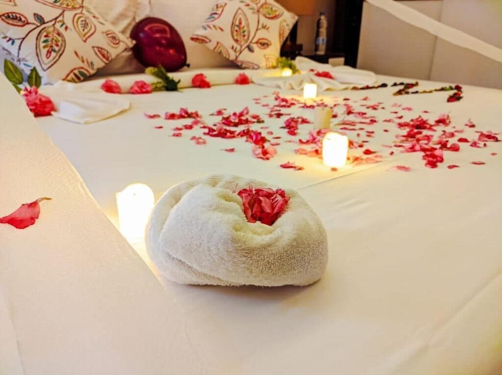 A bed with rose petals and candles, a decorative pillow, and a white towel with a red flower.