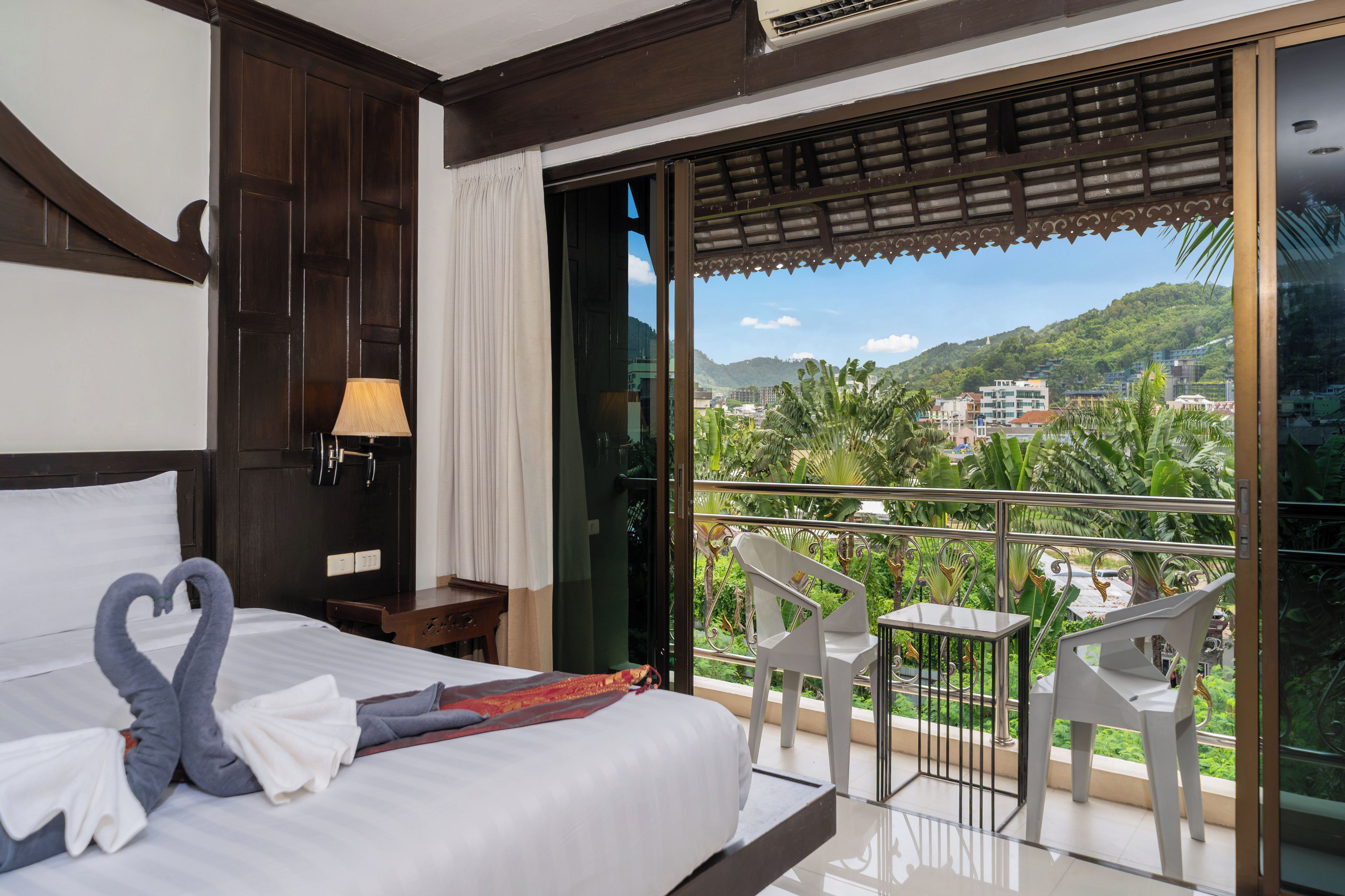 A hotel room with a bed, a nightstand, a lamp, a balcony with chairs, and a view of the city.