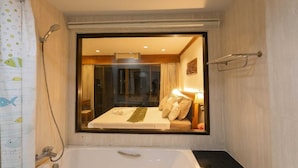 A bathroom with a bathtub, a shower, and a window with a bed visible through it. - Best Venue Hotel (Patong)