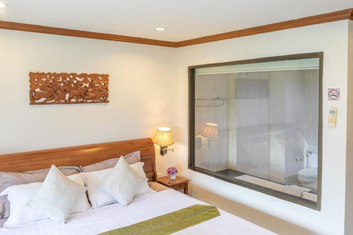 Una habitación de hotel con una cama, una mesita de noche, una lámpara de pared, un ventanal grande y un baño visible a través del ventanal.
