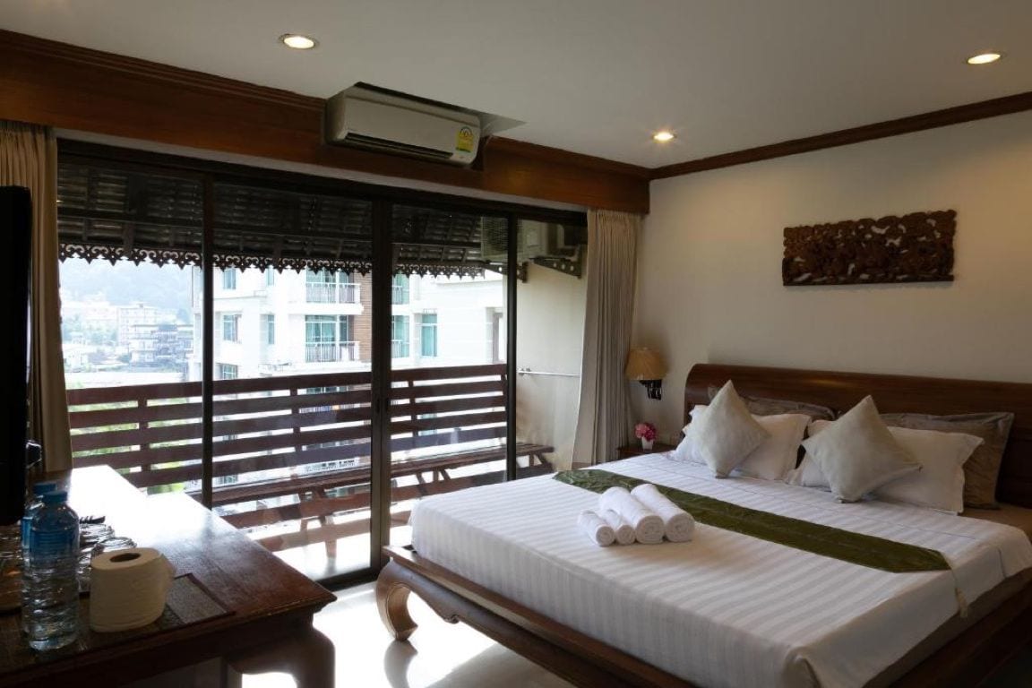 A hotel room with a large bed, a wooden desk, a window with a view, and an air conditioning unit.