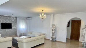 Un salon spacieux avec un grand canapé blanc, une table basse et un lustre. On aperçoit une statue sur le sol et une porte au fond.