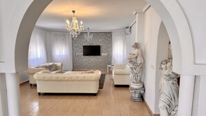 Un salon spacieux avec un grand canapé beige, un canapé plus petit, un téléviseur fixé au mur et un lustre.