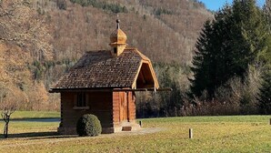 Une petite cabane en bois, dotée d’un toit en pente et d’une cheminée, entourée d’un champ herbeux et bordée, au loin, par une colline boisée.