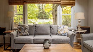 A living room with a grey sofa, two lamps, a wooden coffee table, and a view of trees through large windows. - 1000 FT to Assn Beach, Cute Cottage, Outdoor Dining (Lakeside)