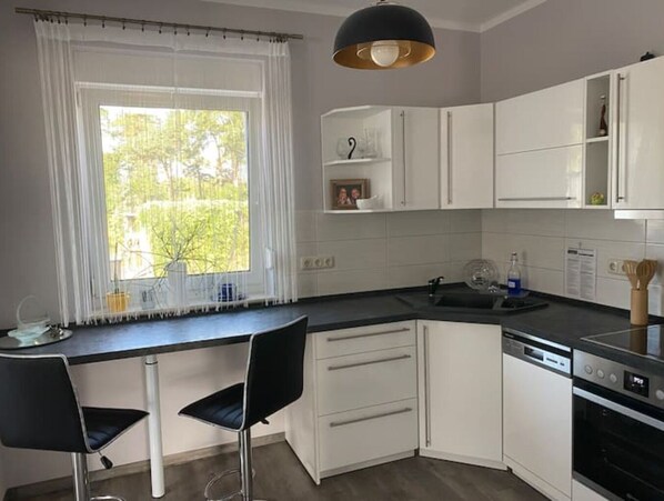 A modern kitchen with white cabinets, a black countertop, a built-in dishwasher, and a large window with white curtains.