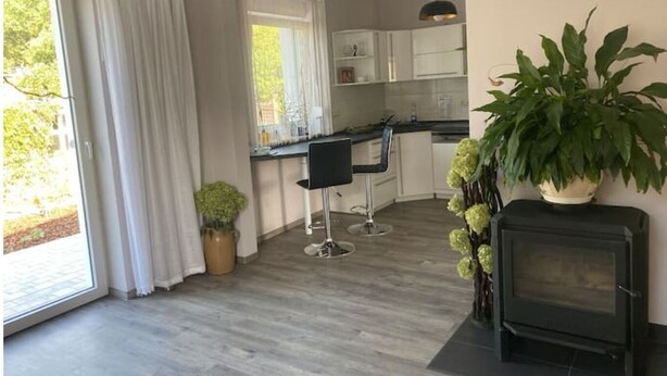A modern kitchen with white cabinets, a black countertop, and a built-in stove. There is a dining area with two chairs and a table, and a large window with white curtains. - Ferienhaus Albertus (Cottbus)