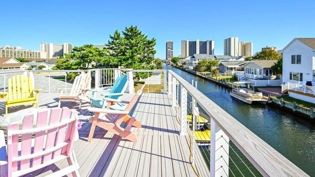 A wooden deck with Adirondack chairs, a view of a canal, and buildings in the background. - Three Bedroom Waterfront Home with a Rooftop Deck (Ocean City)