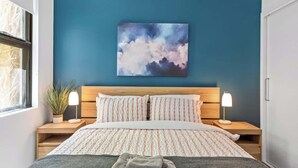 Un dormitorio con cama, dos mesitas de noche, una mesita auxiliar, un cuadro colgado en la pared y una ventana.