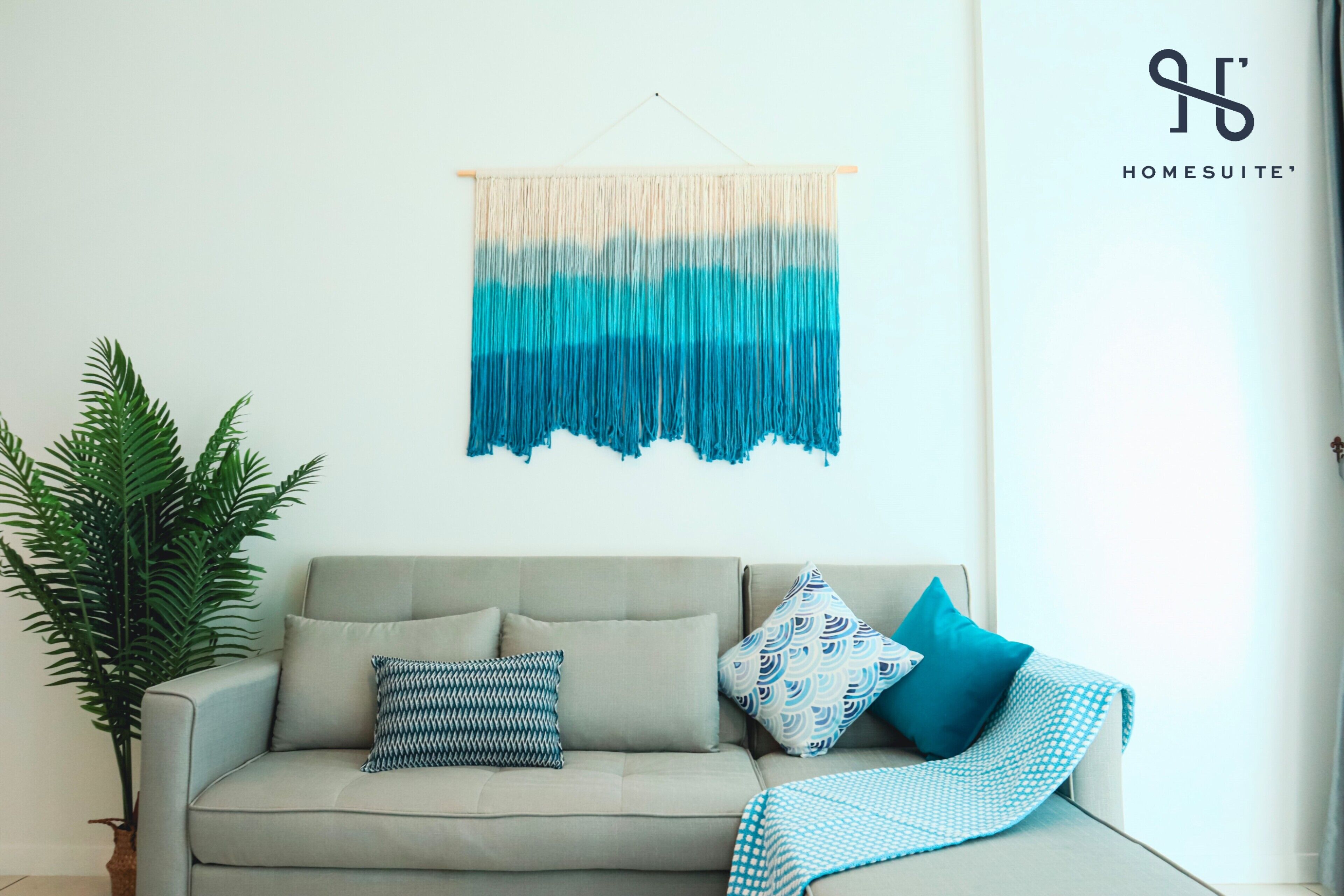 A living room with a sofa, patterned cushions, a blue and white blanket, a hanging macrame wall hanging, and a potted plant.