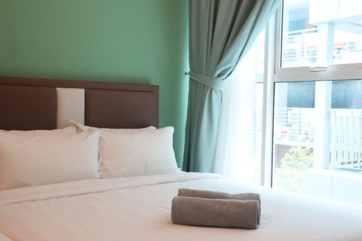 Een hotelkamer met een bed, witte kussens, een bruin hoofdeinde en een raam met gordijnen.