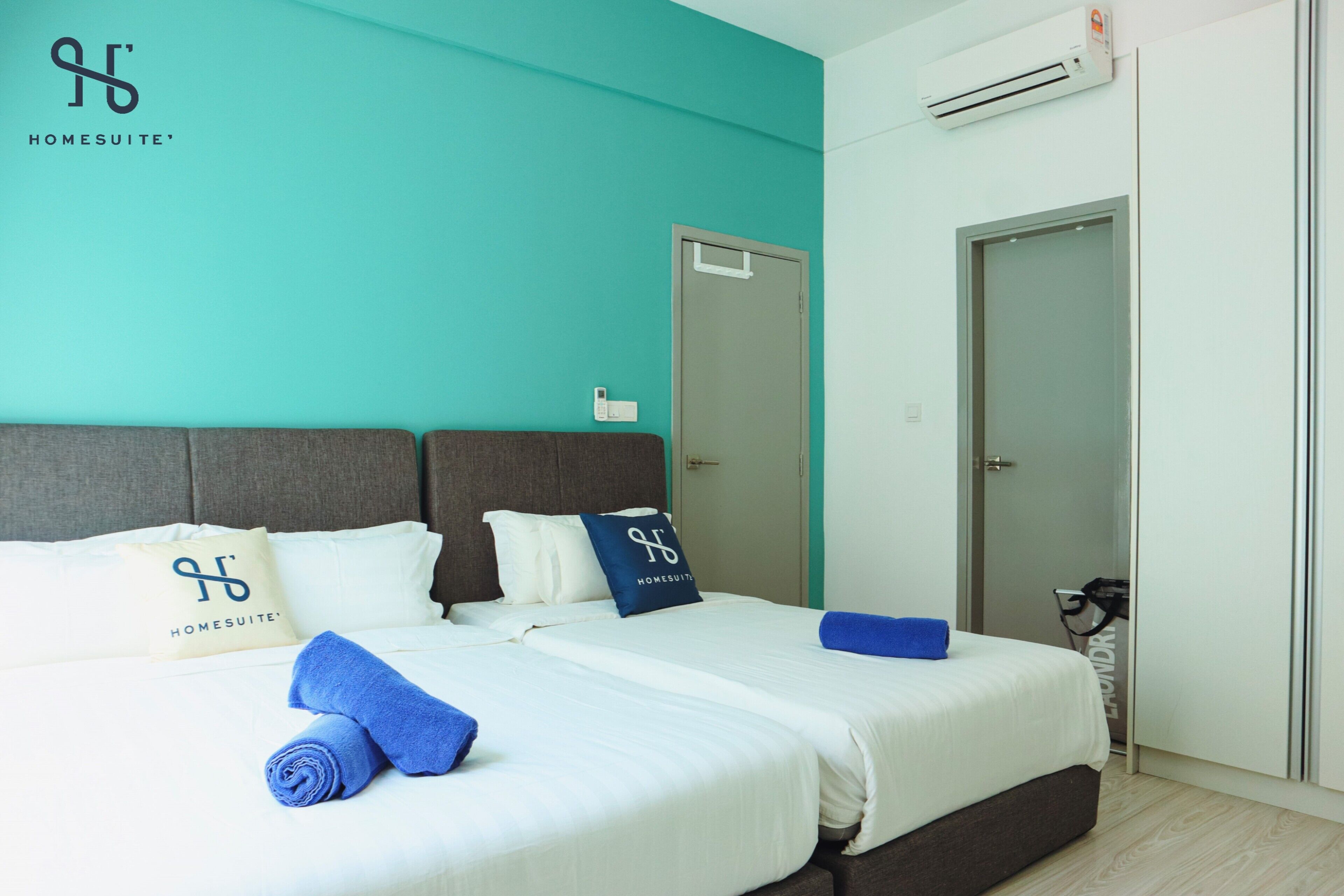 A hotel room with a teal accent wall, a grey headboard, and a white bed with blue towels and pillows.