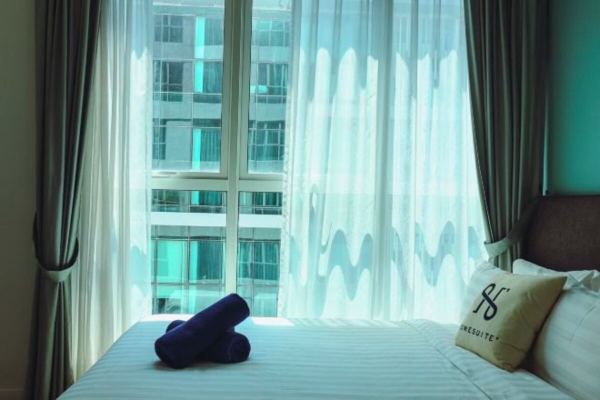 Een hotelkamer met een bed, een kussen met een logo en uitzicht op een gebouw door dunne gordijnen.