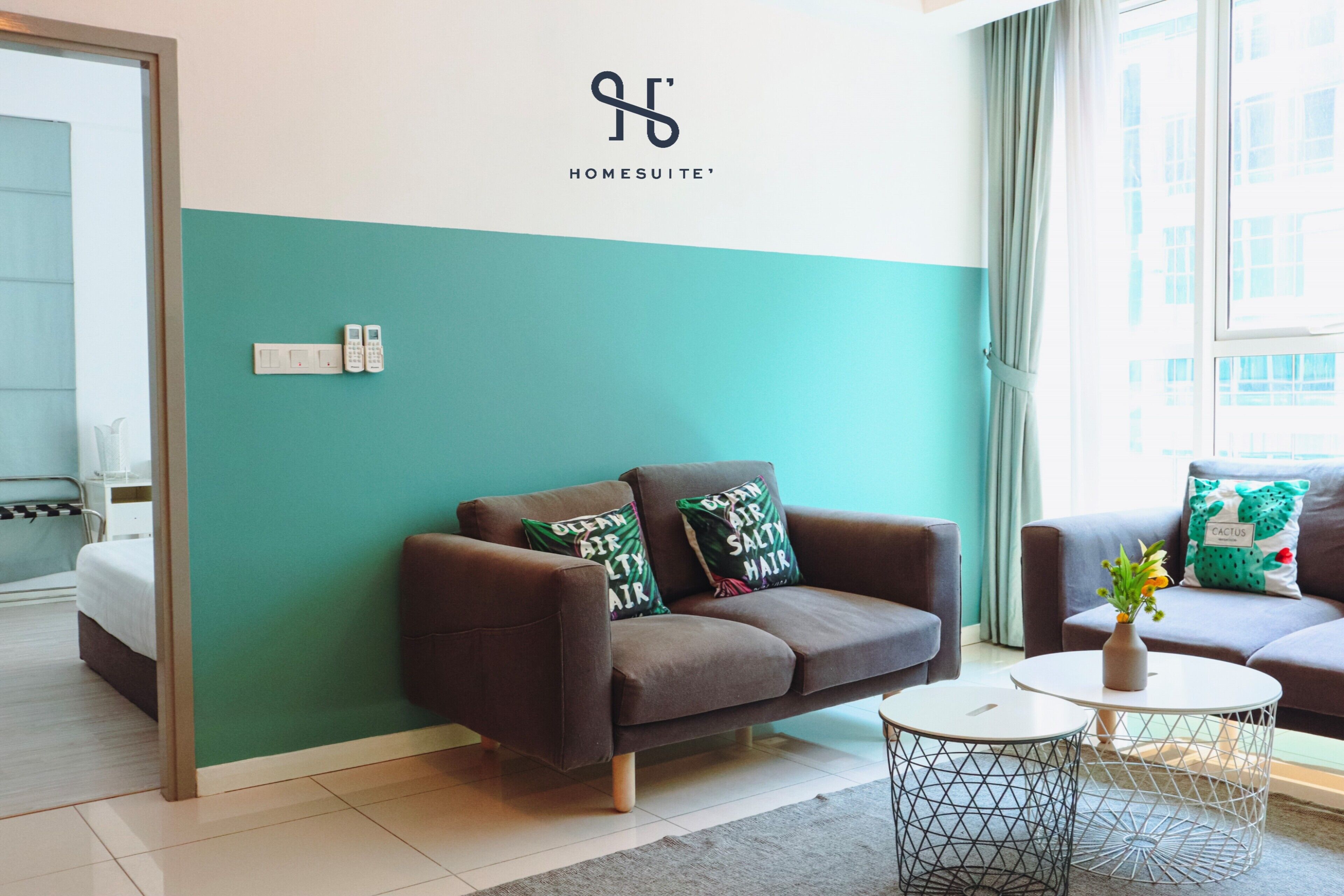 Une chambre d’hôtel moderne avec des murs de couleur turquoise et blanche, un canapé marron, deux petites tables rondes et un lit en arrière-plan.