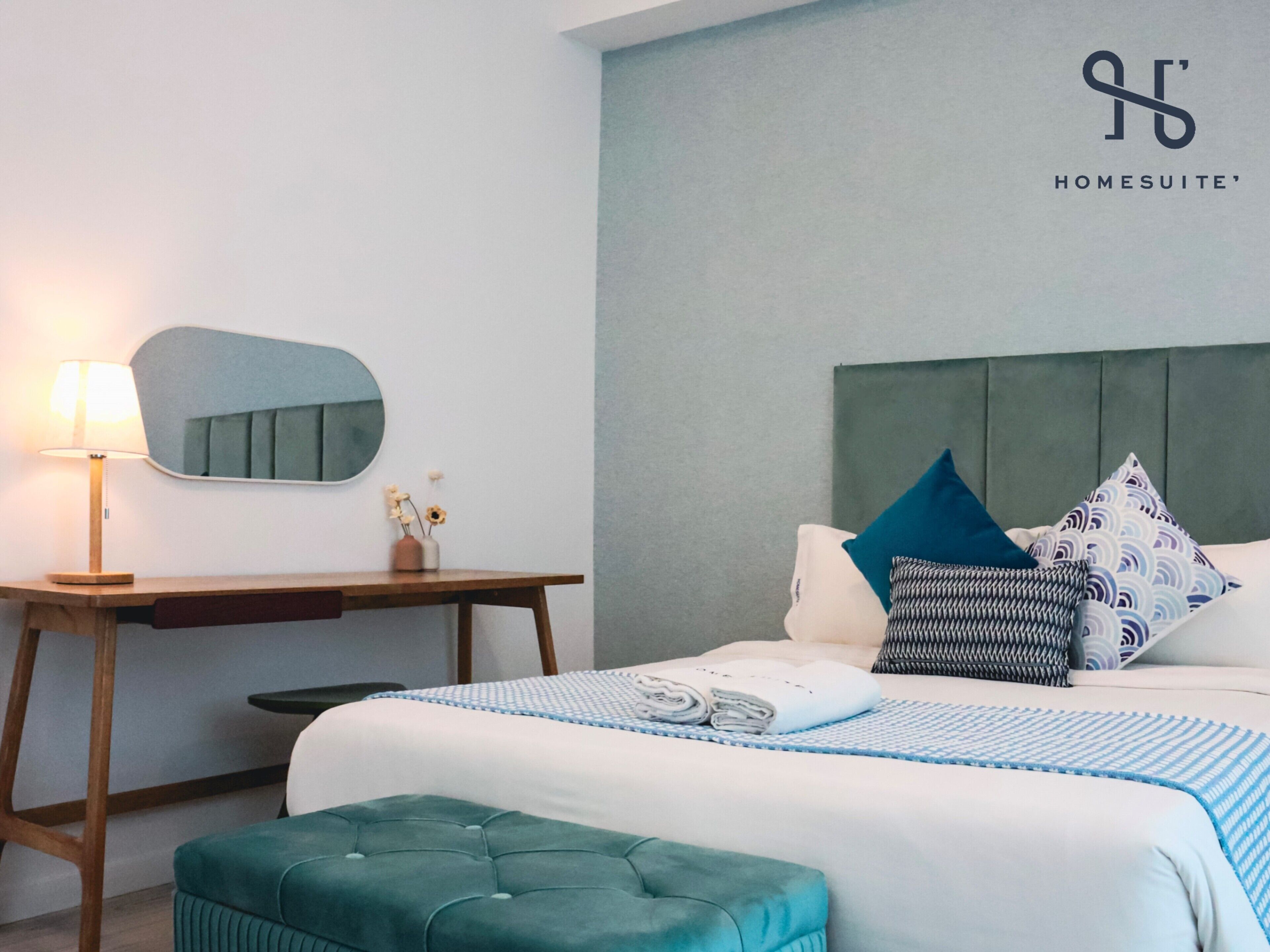 A hotel room with a bed, a desk, a mirror, and a green ottoman.