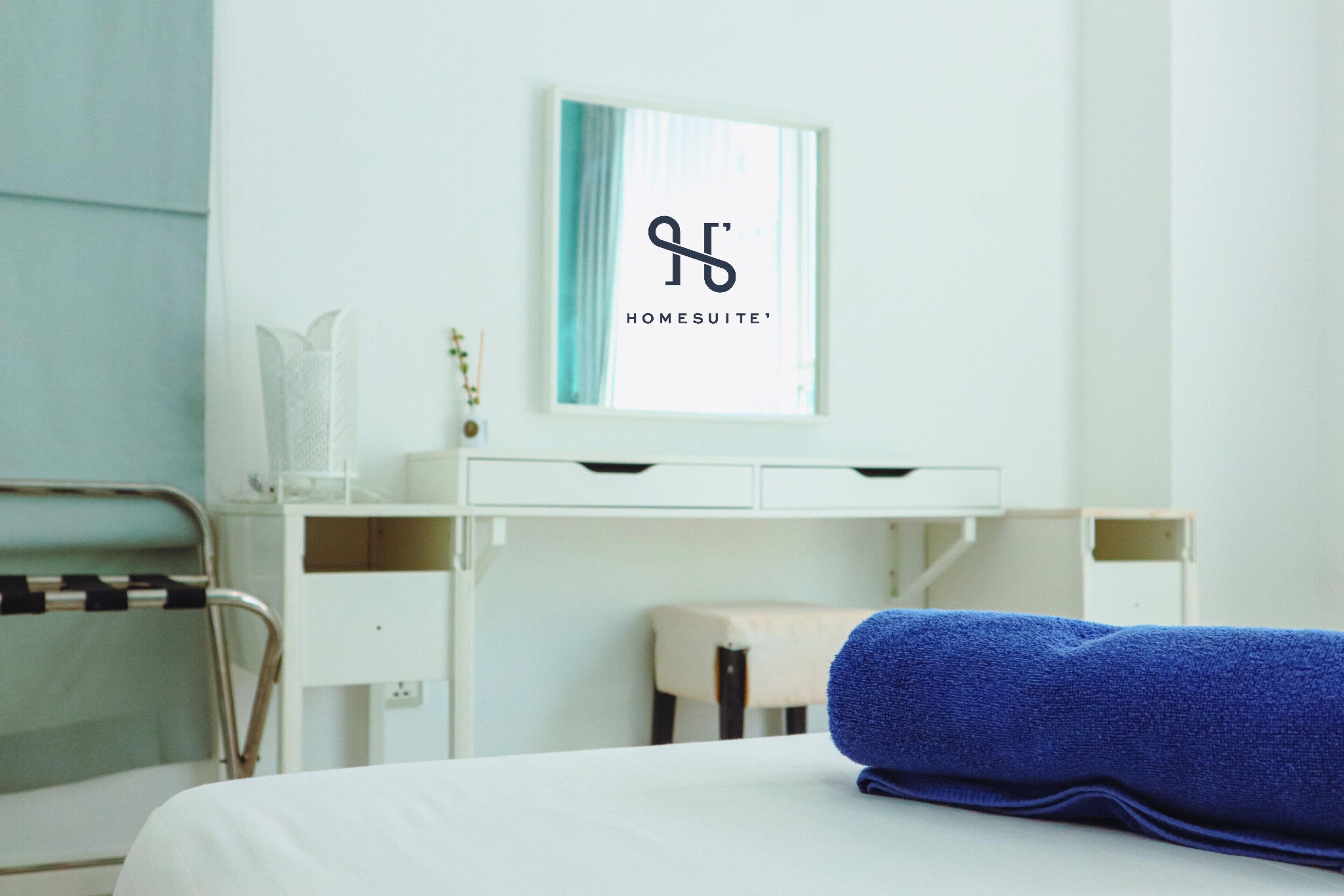 A hotel room with a bed, a desk with a computer, a chair, and a blue towel on the bed.