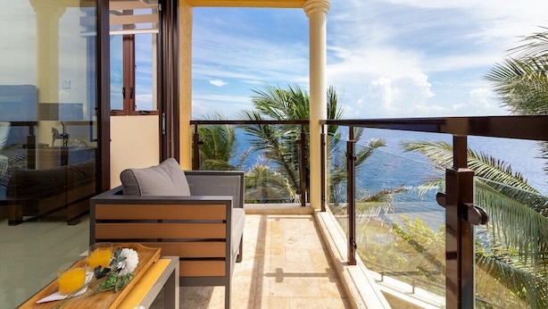 Un balcon avec un canapé et un plateau de jus d’orange, offrant une vue sur la côte, avec des palmiers et un ciel dégagé.