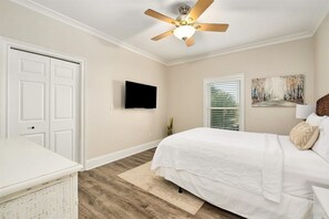 A bedroom with a bed, a ceiling fan, a television, a door, and a window with blinds.
