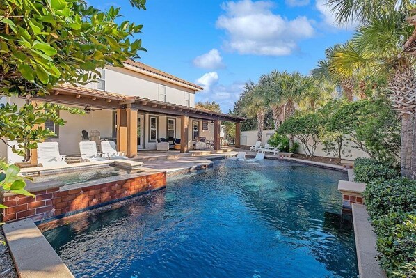 A swimming pool with lounge chairs, a covered patio, and a house with a tiled roof.