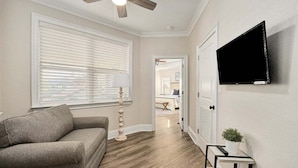 A living room with a sofa, a ceiling fan, a television mounted on the wall, and a hallway leading to another room.