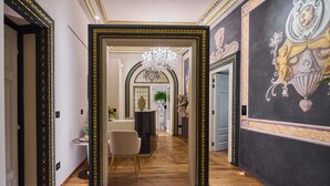 A hallway with a grand chandelier, ornate black and gold frames, and a marble floor. - Duomo Luxury Florence (Florence)