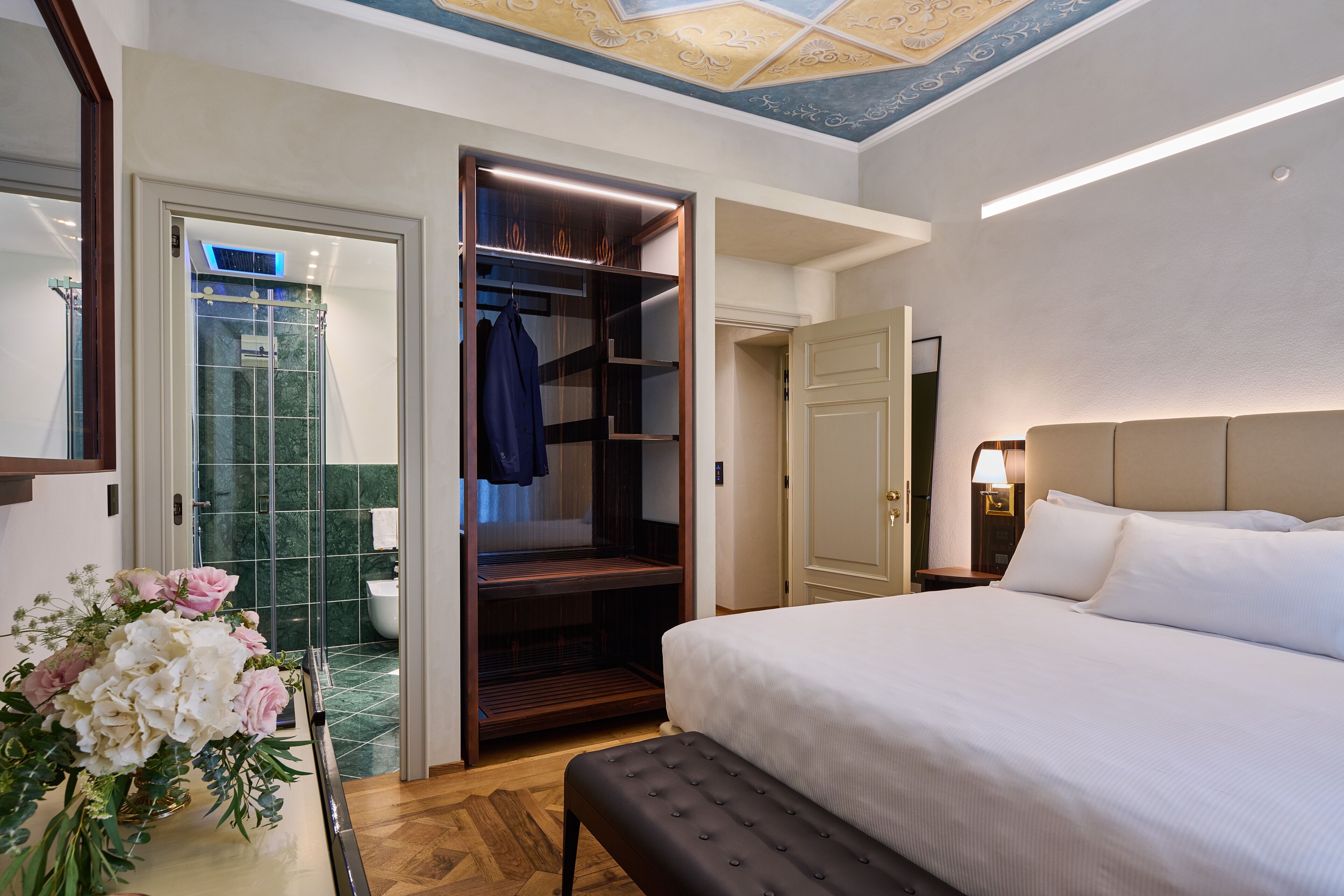 A hotel room with a large bed, a walk-in closet, a bathroom with a glass shower, and a ceiling with a decorative pattern.
