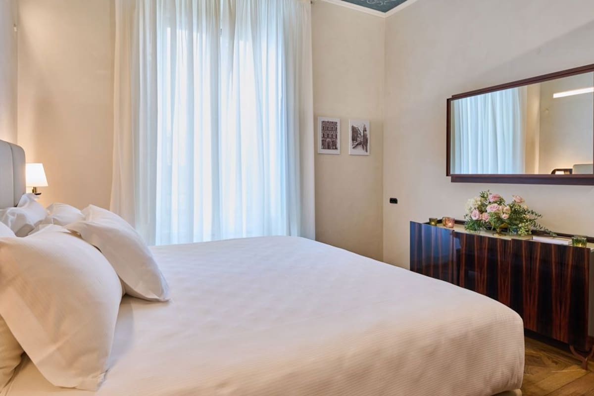Habitación de hotel con una cama grande, una mesita de noche con lámpara, un espejo y un pequeño aparador con flores.