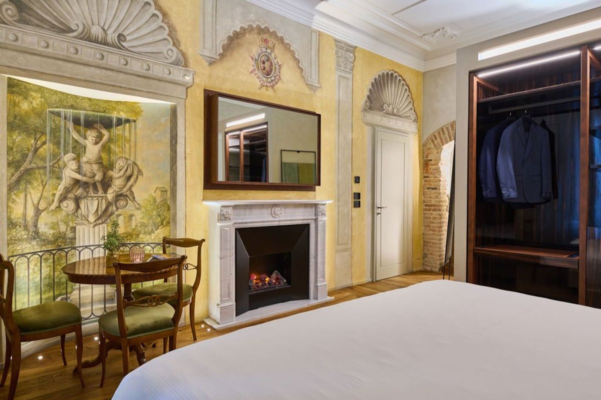Une chambre d’hôtel avec une cheminée, un grand lit, une table à manger avec des chaises, une armoire et une fresque murale.