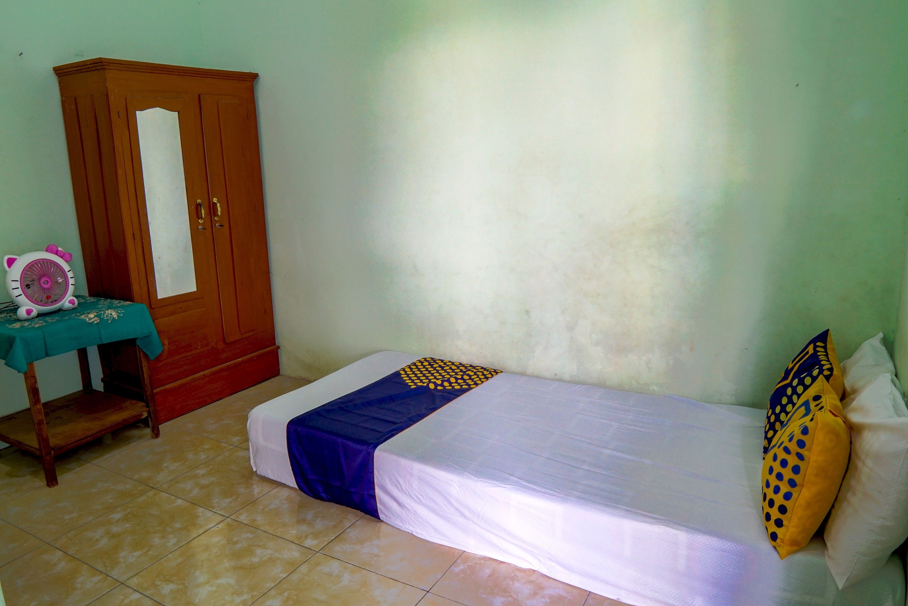 Quarto pequeno com uma cama de solteiro, um guarda-roupa de madeira e uma mesinha com um leque rosa.