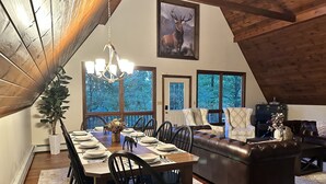 A dining area with a long wooden table set for six, surrounded by black chairs, a chandelier, and a large window offering a view of trees.