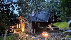 A stone cabin with a wooden deck and a stone chimney, surrounded by trees and a wooden pathway.