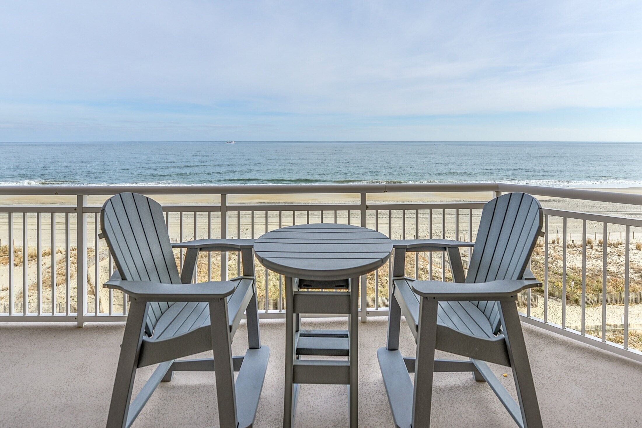 A balcony with a table and two Adirondack chairs overlooking a beach.