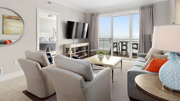 Ein modernes Wohnzimmer mit einer Couch, Sesseln, einem Couchtisch und einem Fernseher. Ein Balkon bietet Blick auf den Himmel.
