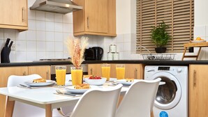 A modern kitchen with white cabinets, a built-in washing machine, and a dining area with white chairs and a table set for breakfast. - Perfect for Family & Corporates - FREE Parking - Contact us for better rates! (Headingley)