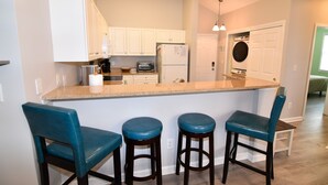 A kitchen with white cabinets, a granite countertop, a built-in oven, and a microwave. There are three blue bar stools and a white door with a circular window.