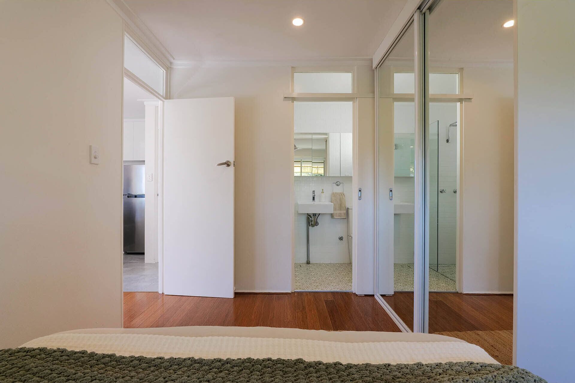 Une chambre moderne avec un grand lit, une porte-fenêtre coulissante donnant sur une salle de bain et une cuisine visible à travers une porte ouverte.