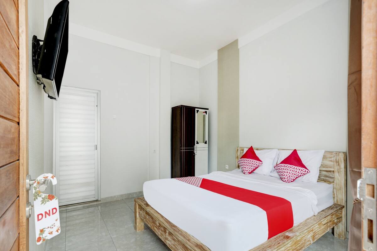 Une chambre avec un lit en bois, du linge de lit blanc et des oreillers rouges. On y trouve une armoire et un téléviseur fixé au mur.