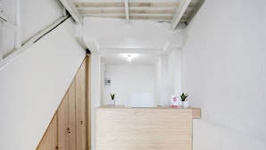 A reception desk in a hallway with a wooden staircase on the left and a white wall with a ceiling light. - OYO Flagship 90756 Melasti Hills Room & Cafe (Ungasan)