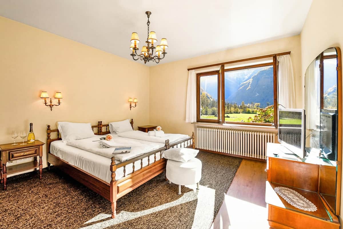 A bedroom with a wooden bed, a chandelier, a large window with a mountain view, a bedside table with glasses, and a TV.