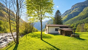 A house with a stone exterior, surrounded by a grassy area and trees, with mountains in the background. - Casa Mima Chalets (Sonogno)