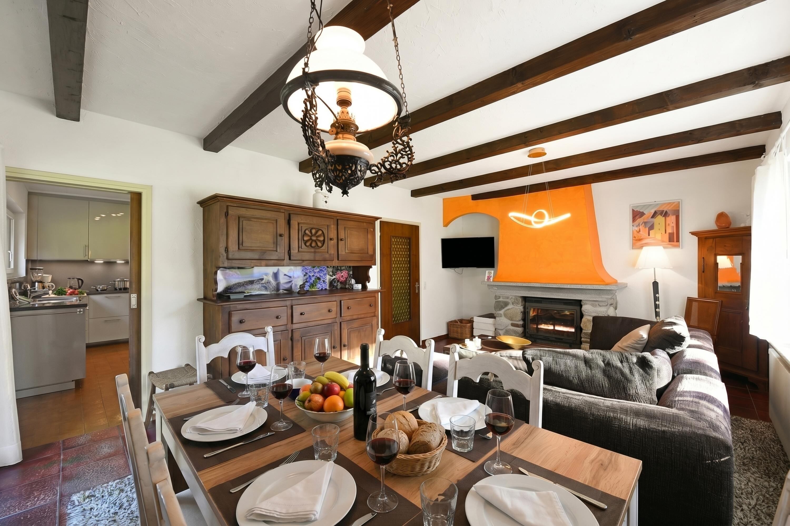 Un coin repas avec une table en bois dressée pour le repas, une cuisine en arrière-plan et un salon avec un canapé et une cheminée.