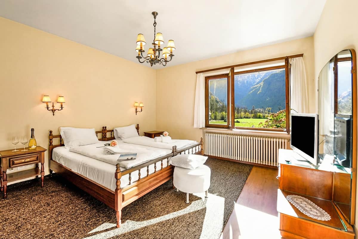 A hotel room with a bed, bedside tables, a TV, a window with a mountain view, and a chandelier.
