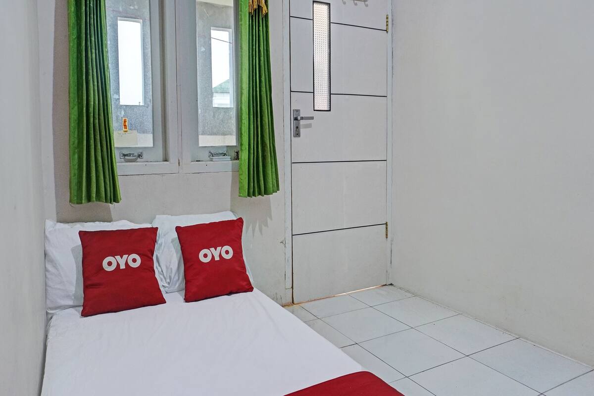 A bed with white bedding and two red pillows with the word "OYO" on them, a window with green curtains, and a white door with a rectangular light panel.