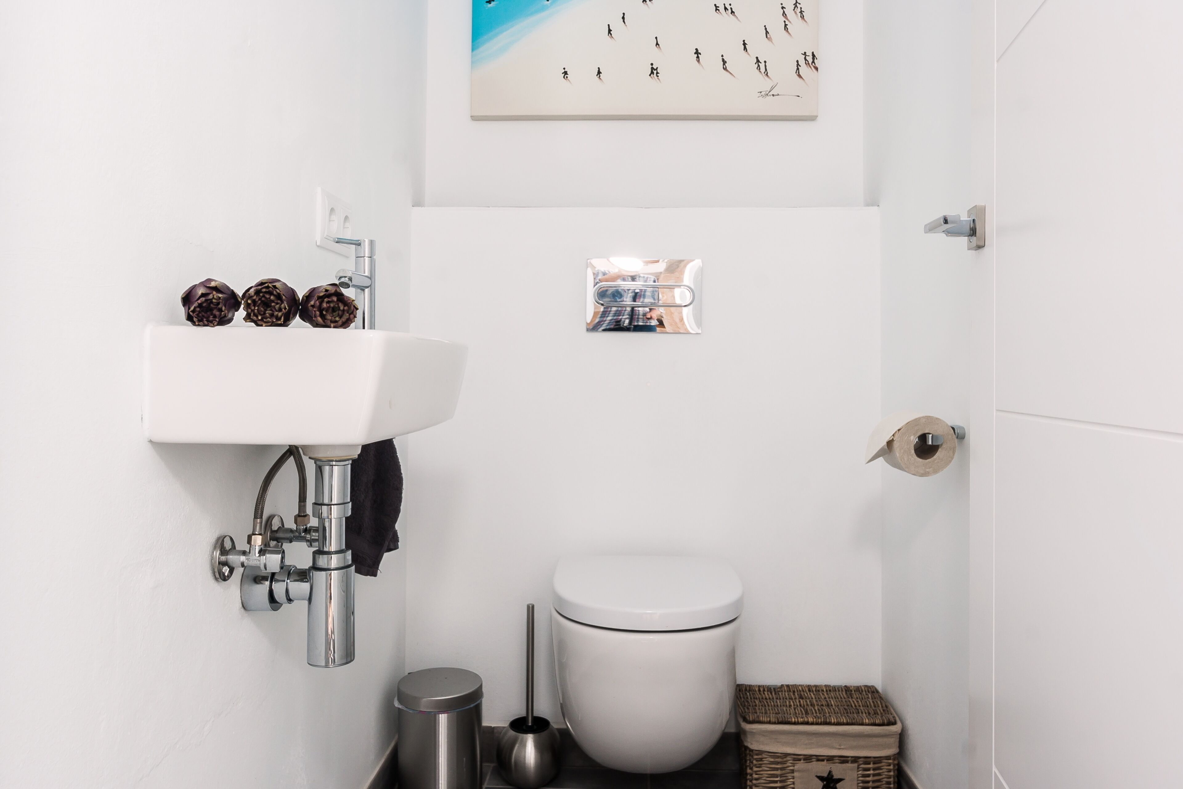 A bathroom with a white sink, toilet, and a small trash bin. There is a picture frame on the wall and a basket on the floor.