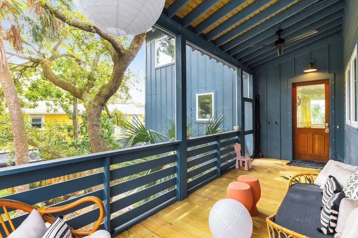 Een overdekt terras met houten vloer, een rieten stoel, een comfortabele bank en een klein tafeltje.